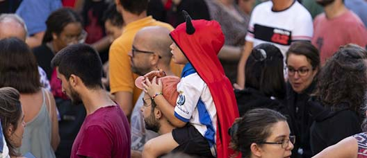 Un niño que lleva una capucha de demonio, subido a hombros de un hombre entre el público del pregón de las fiestas de Gràcia